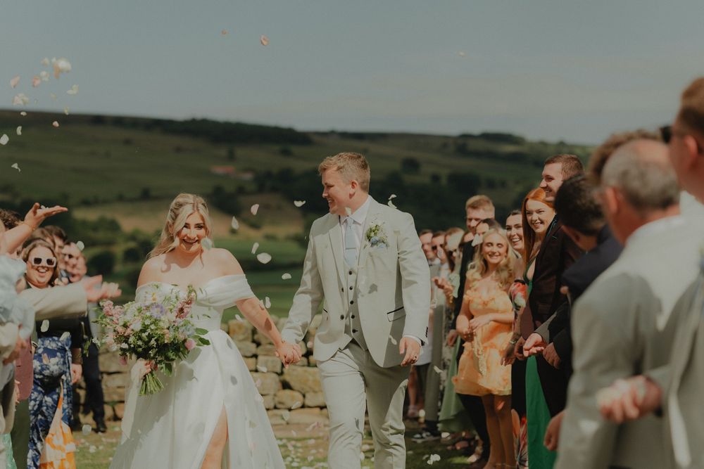 Bride and groom walk through confetti shower with guests cheering outdoors at countryside wedding venue.