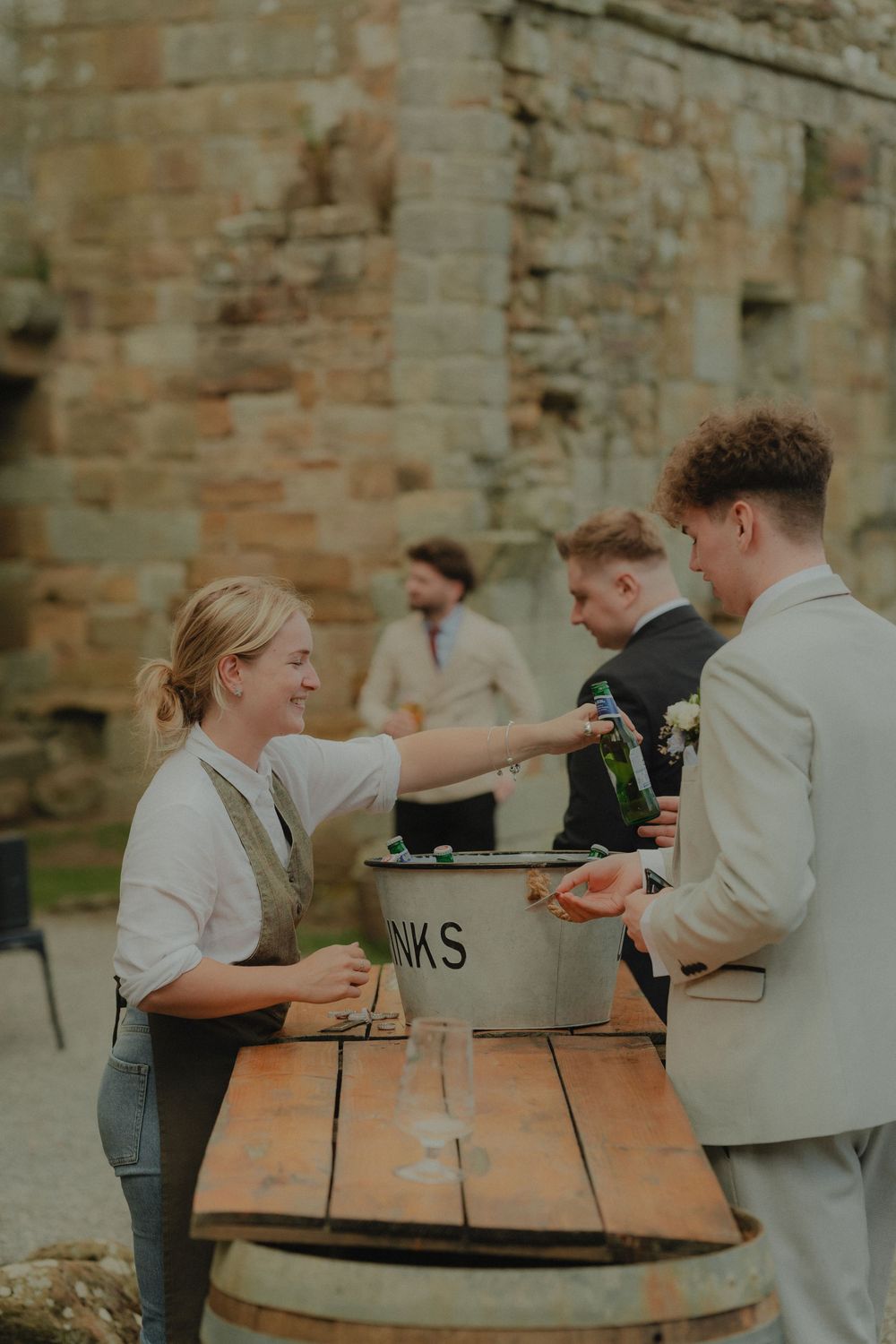 Smiling female bartender serves drinks from a metal bucket on a wooden barrel at an outdoor wedding reception.