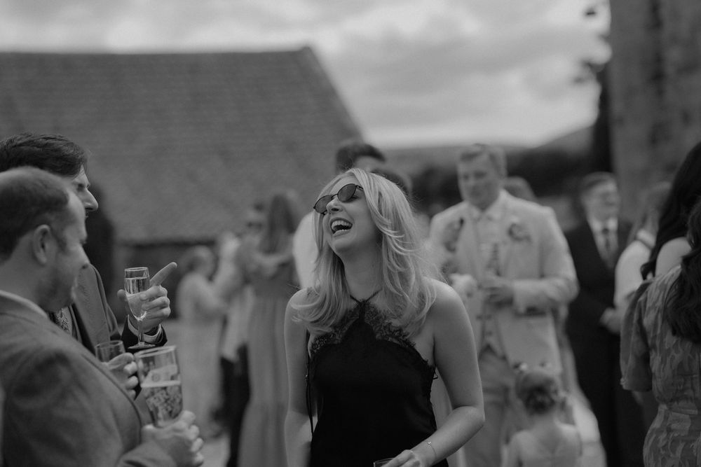 Black and white photo of a laughing blonde woman in a black dress socializing at an outdoor wedding reception.
