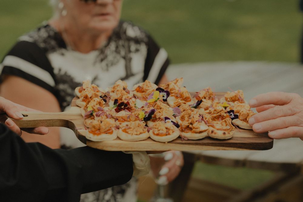 A wooden serving board filled with elegant bite-sized appetizers topped with colorful garnishes held by two people outdoors.