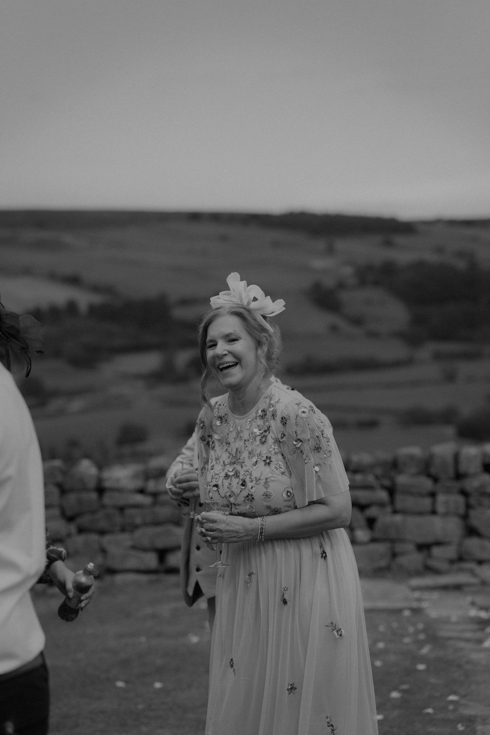 Joyful wedding guest laughing outdoors in floral dress and fascinator with rolling hills in black and white.