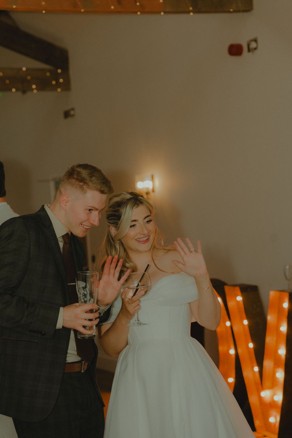 Bride in white gown and groom in dark suit wave and smile holding champagne glasses at wedding reception.