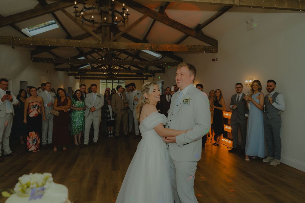 Bride and groom share their first dance in a rustic barn venue surrounded by wedding guests clapping and celebrating.