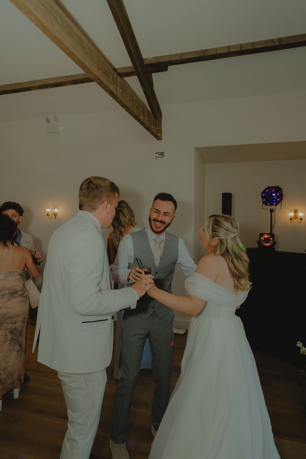 Bride and groom laughing and dancing with a guest at their wedding reception in a rustic venue with wooden beams.