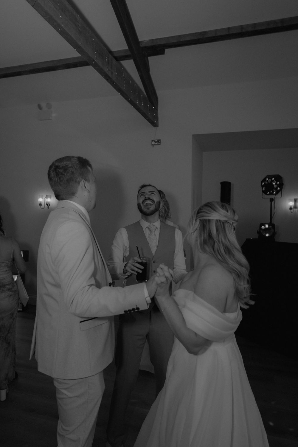 Black and white photo of bride and groom dancing joyfully with a smiling guest at a rustic barn wedding reception.
