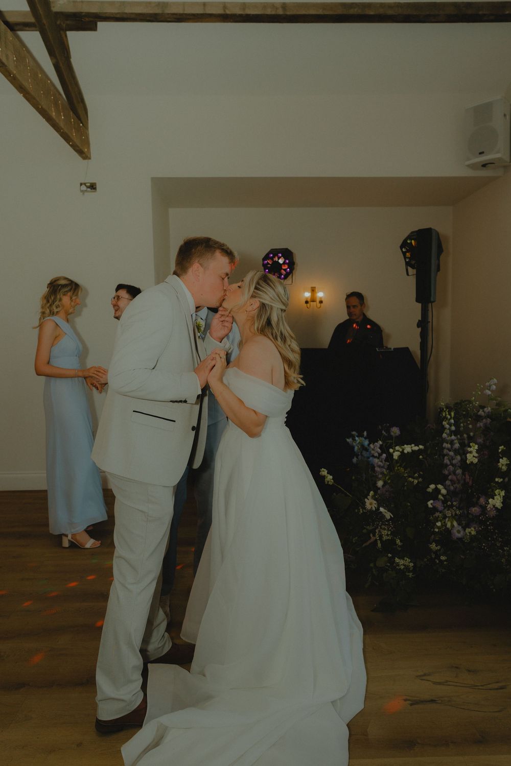 Bride and groom share a first dance kiss at their wedding reception with guests and DJ in the background.