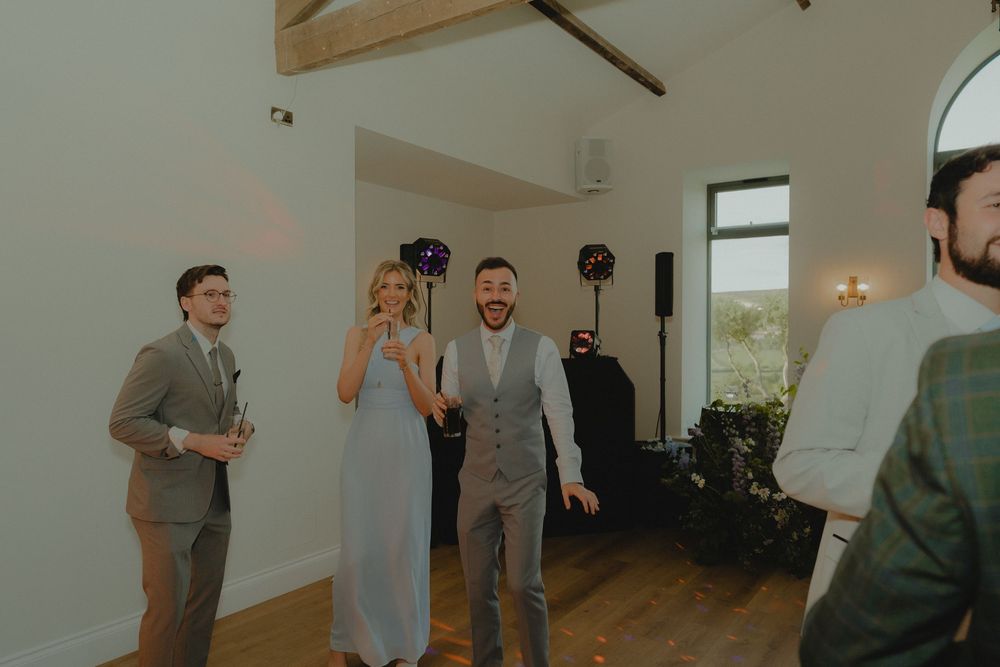 Guests laughing and dancing at a rustic barn wedding reception with wooden beams and warm lighting.