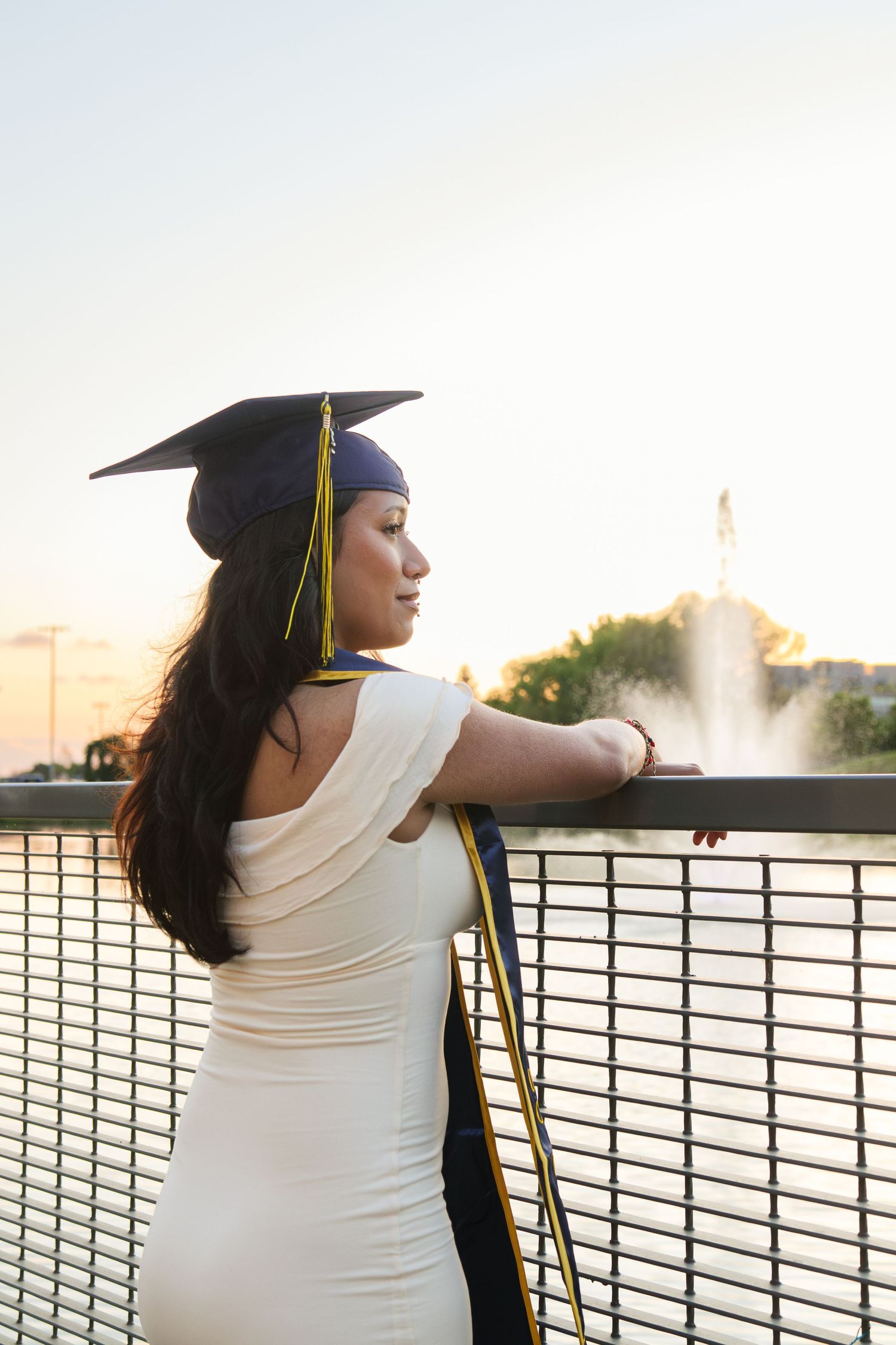 Graduate in cap and gown leaning on a railing overlooking a fountain at sunset.