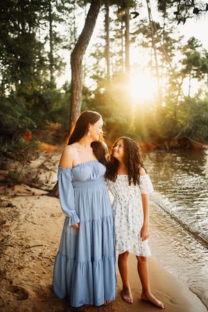 Mother and daughter standing on a beach.  They are looking at each other lovingly and the sun is setting behind them.