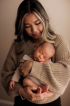 Sleeping newborn baby in beige knit sweater nestles against mother in matching cozy sweater during intimate portrait.