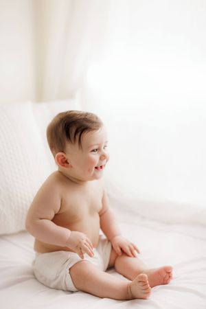 Happy baby in white diaper sits on soft white bedding while smiling and looking to the side.