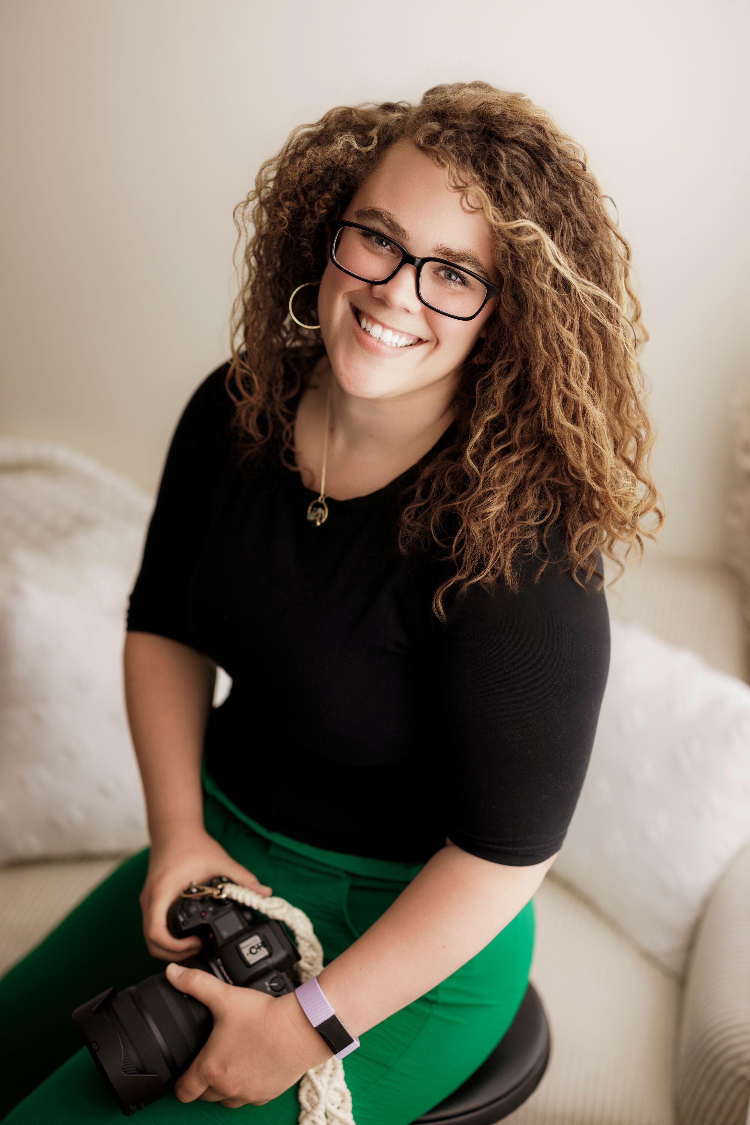 Photographer with curly blonde hair wearing black top and green pants sits on white couch holding professional camera.