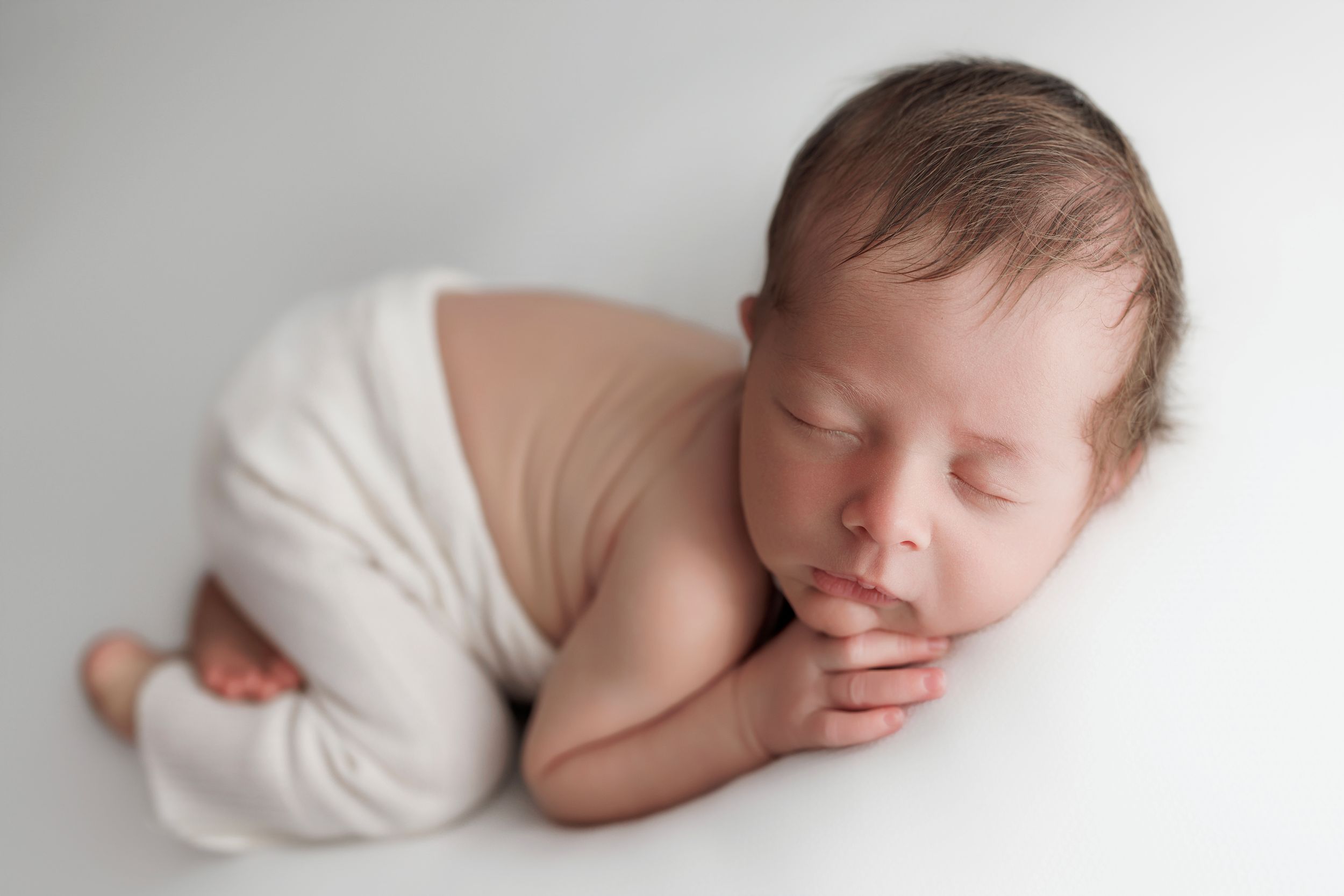 Peaceful sleeping newborn dressed in white wrapping curled up in fetal position on soft backdrop.