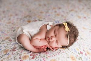 Sleeping newborn in white onesie with yellow bow rests on floral patterned blanket.