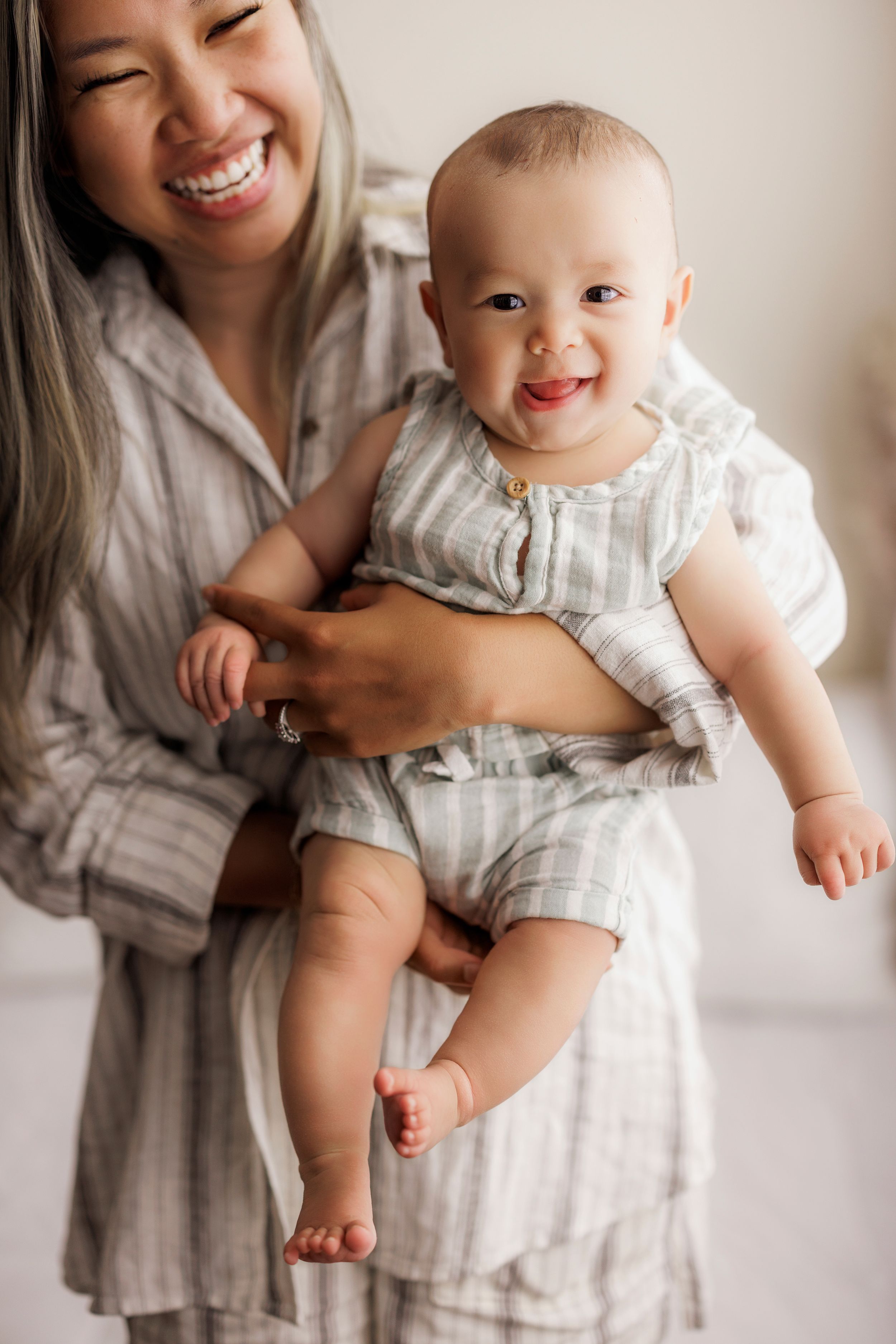 A happy baby in a striped romper is held by someone wearing a light linen dress in a bright interior setting.