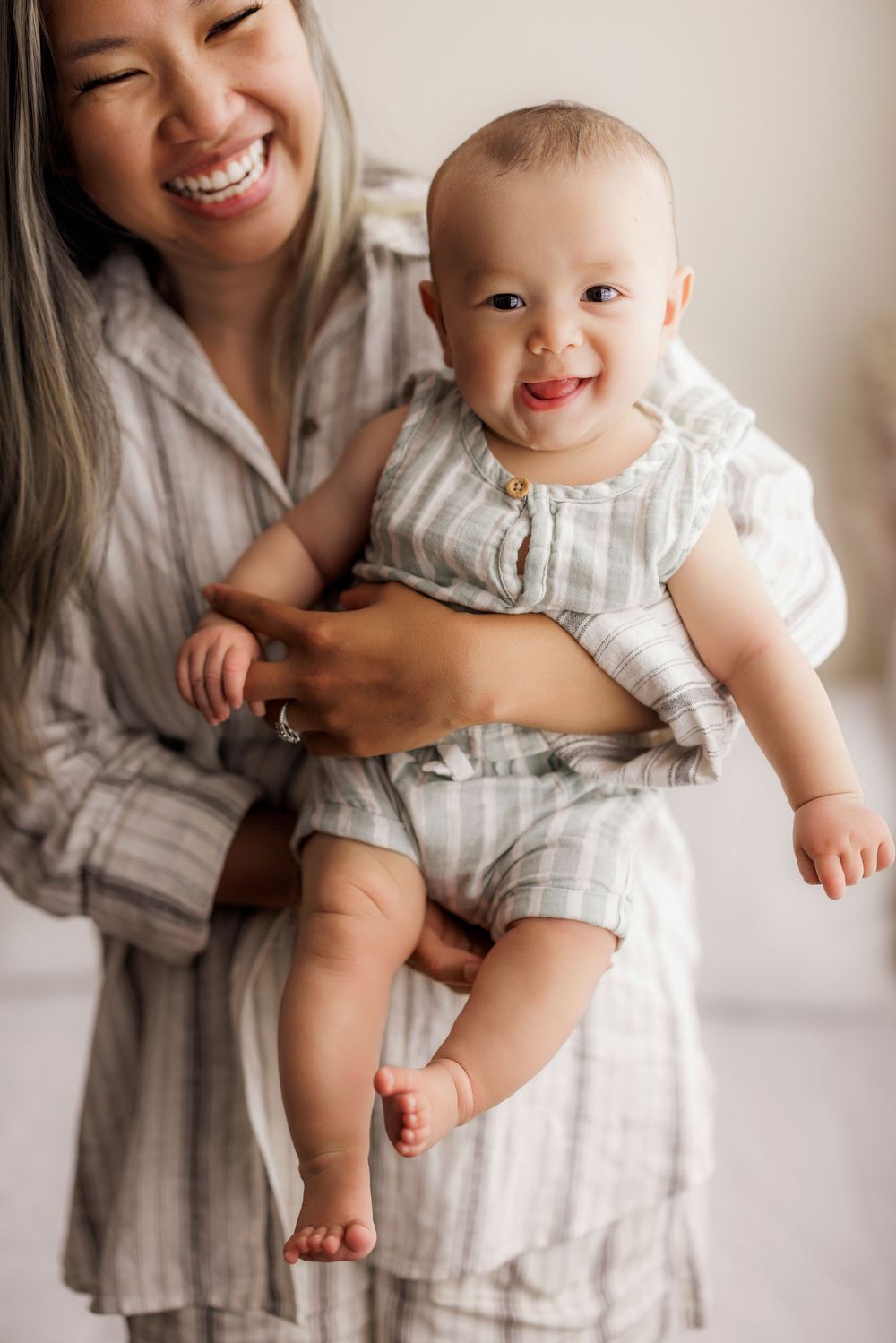A happy baby in a striped romper is held by someone wearing a light linen dress in a bright interior setting.