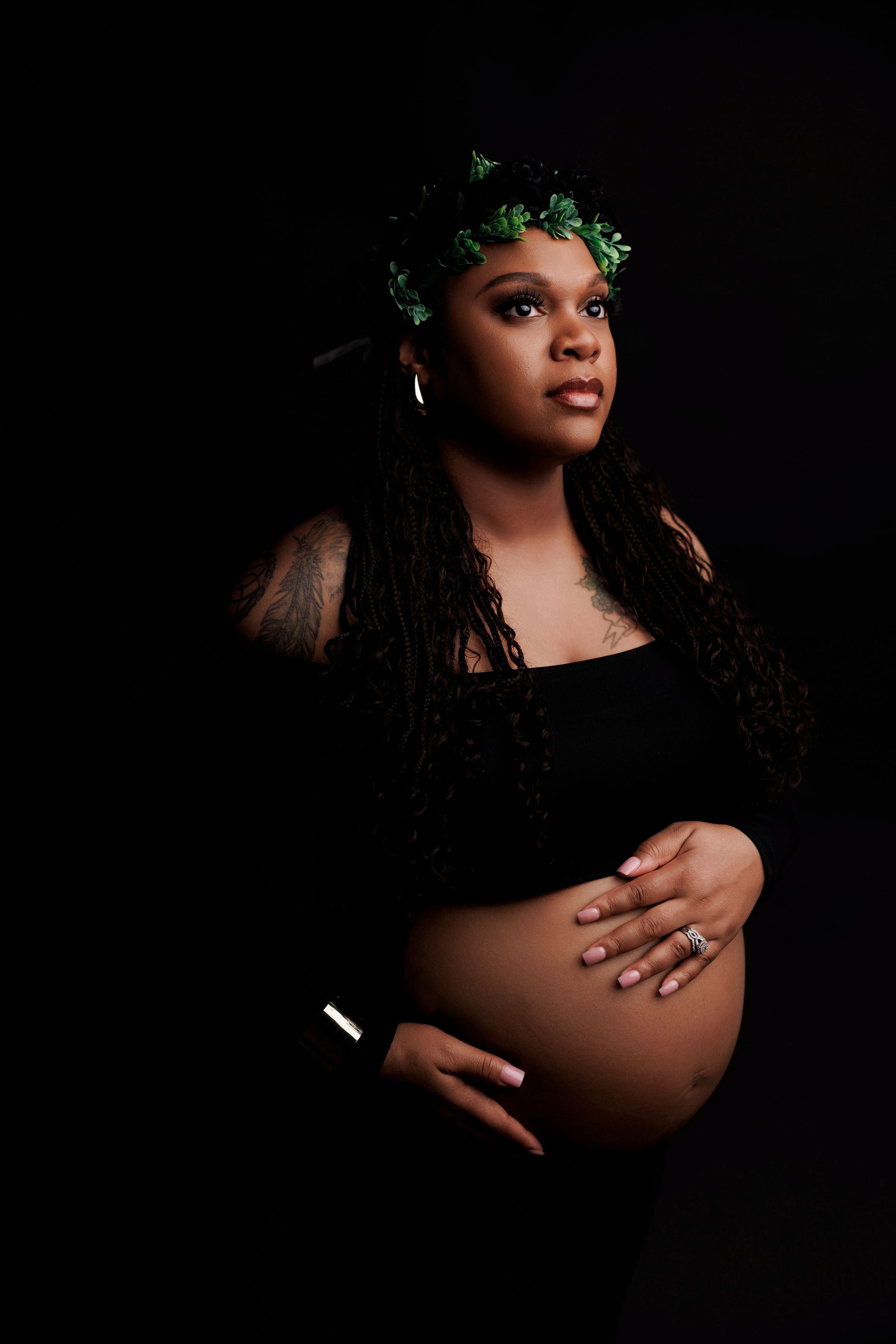 Elegant maternity portrait against dark background with delicate green floral crown and long dark hair.