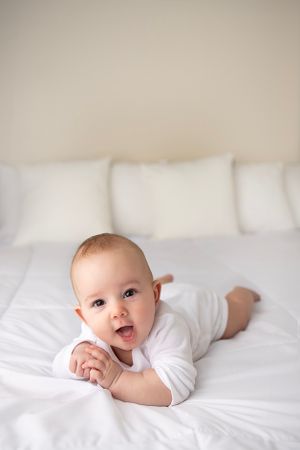 Four month old baby boy on a white bed.  Posed up his elbows smiling at the camera.