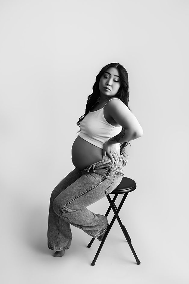 Black and white image of pregnant mother in studio sitting on a stool showing her belly.