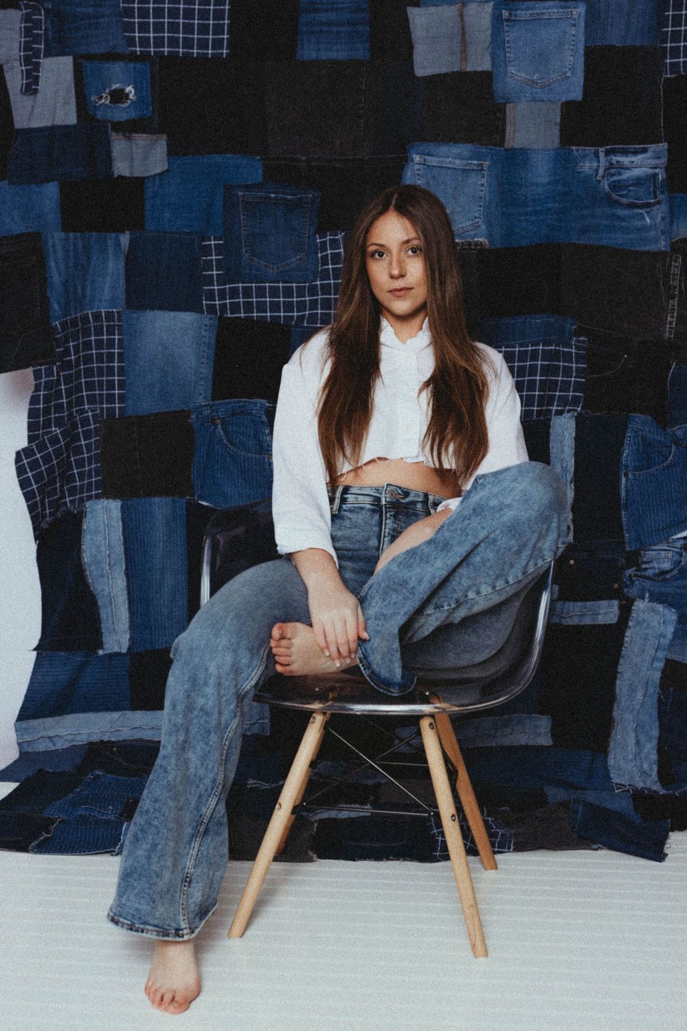 Young woman in white crop top and wide-leg jeans sits barefoot on modern chair against denim patchwork backdrop.