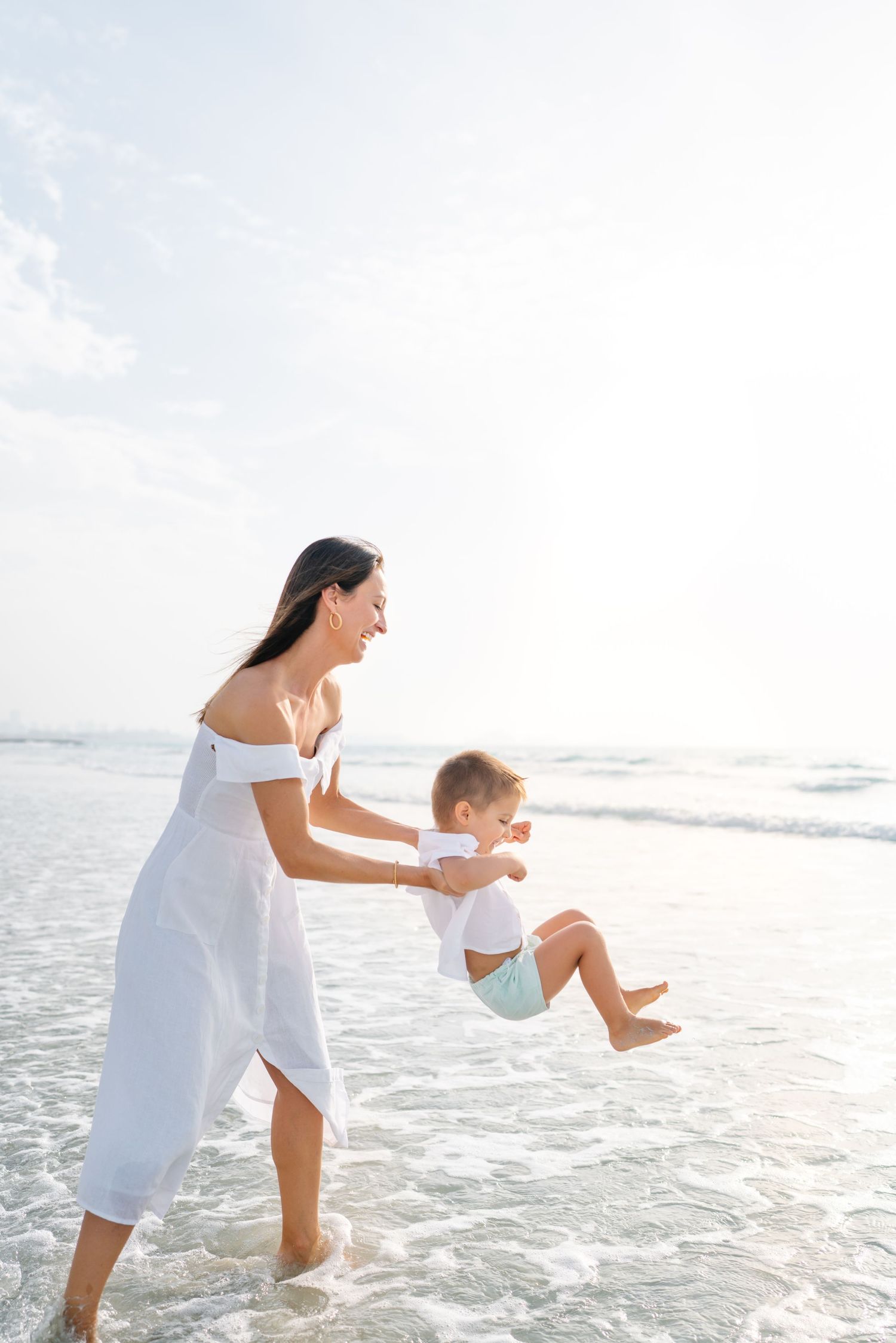 Mother in white dress swinging child playfully at beach during sunset.