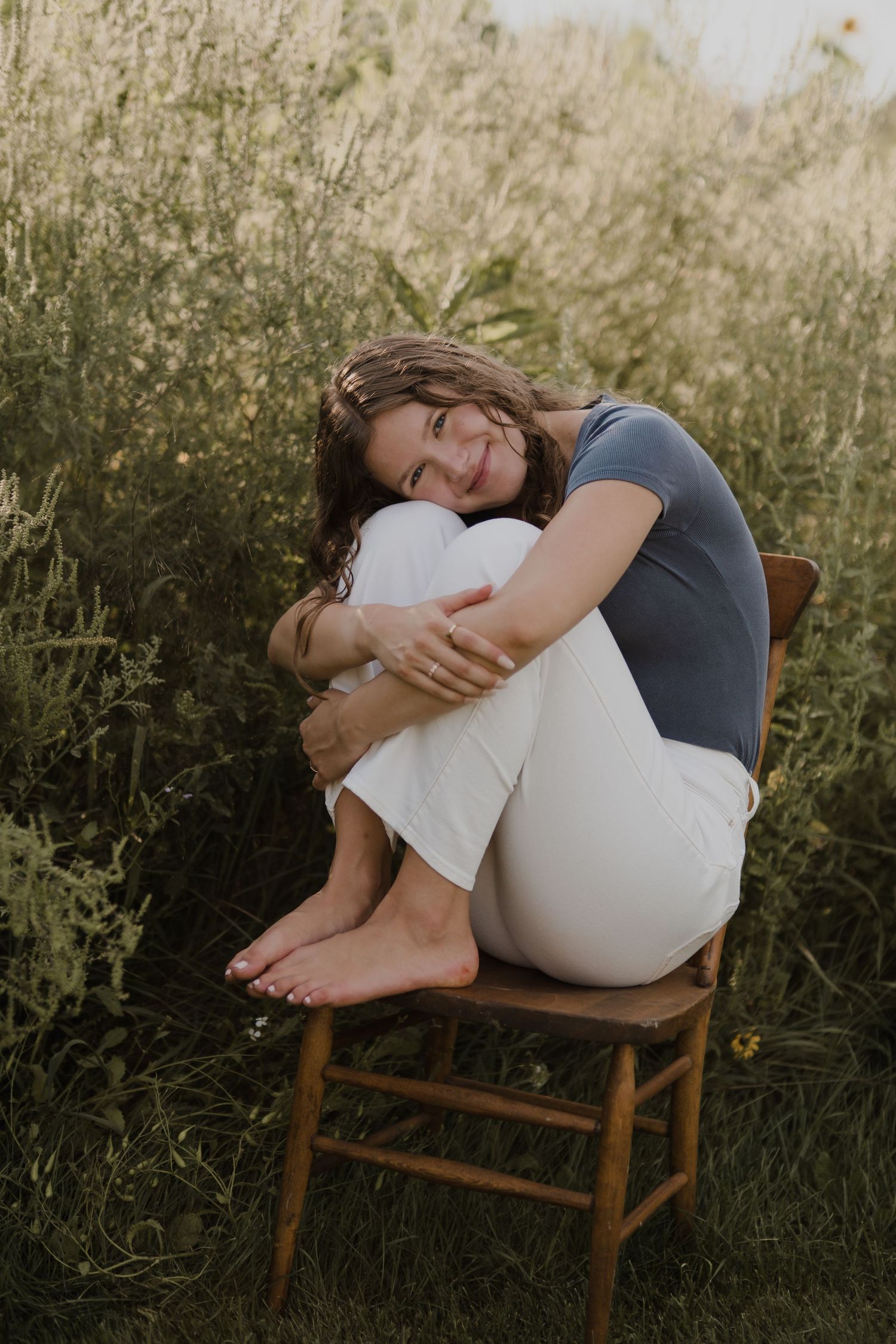 Artistic portrait on a wooden chair surrounded by tall grass in golden sunlight.