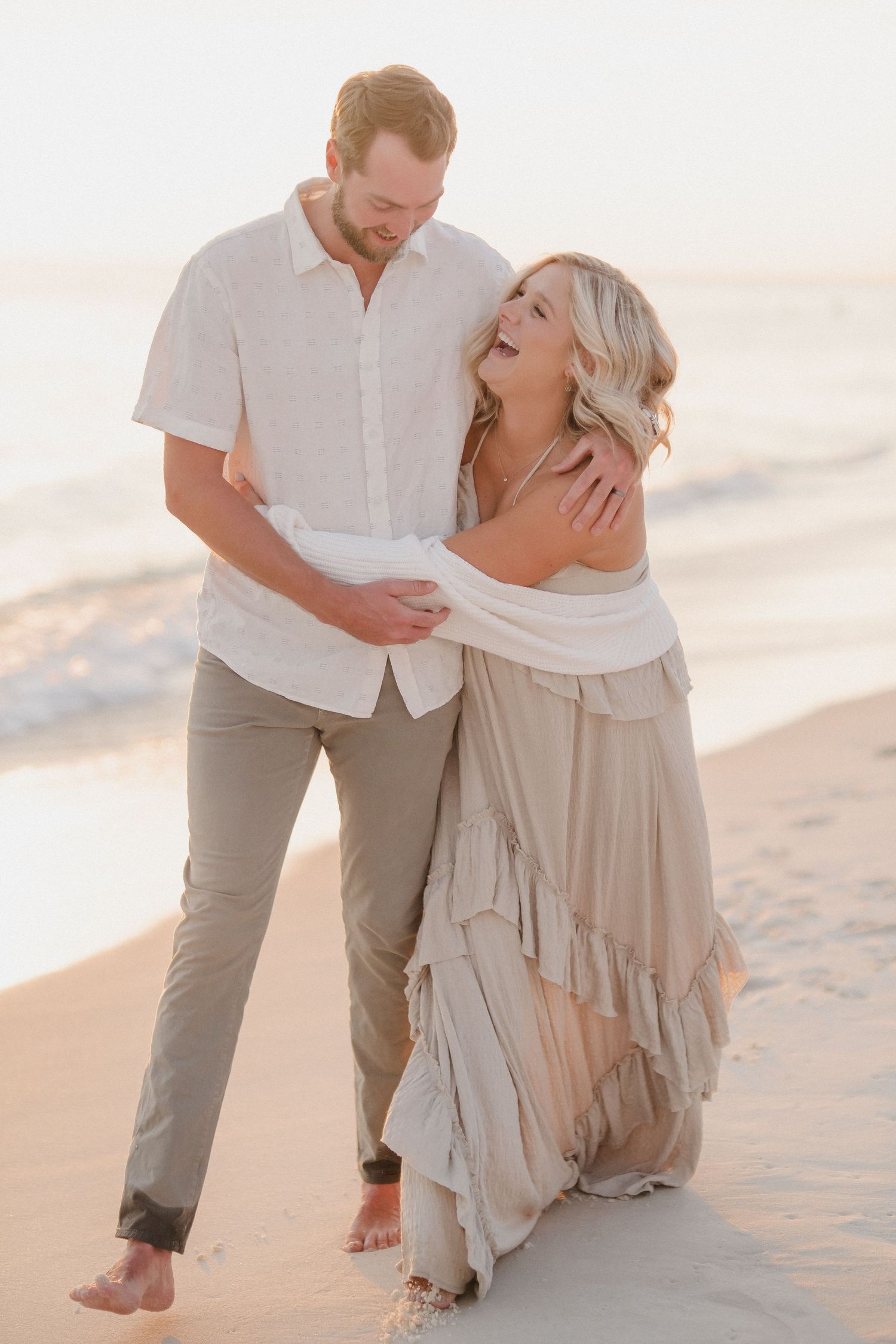 Romantic couple in white and beige outfits embracing on a sandy beach at sunset.