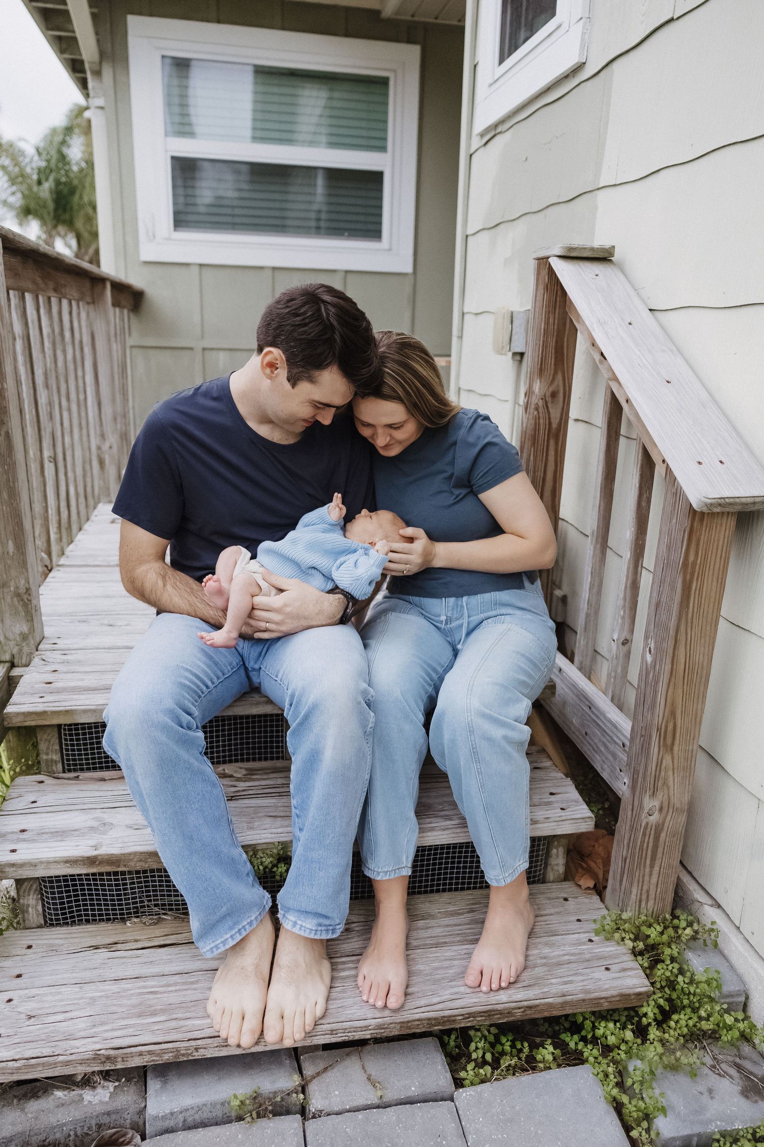 New parents sitting on wooden porch steps cradling newborn baby together in casual denim outfits.