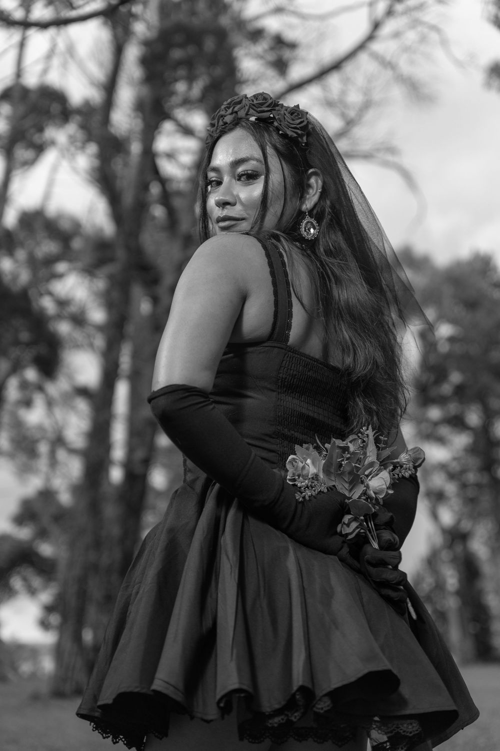 Black and white portrait of person in a dramatic gothic dress and crown posing in a woodland setting.
