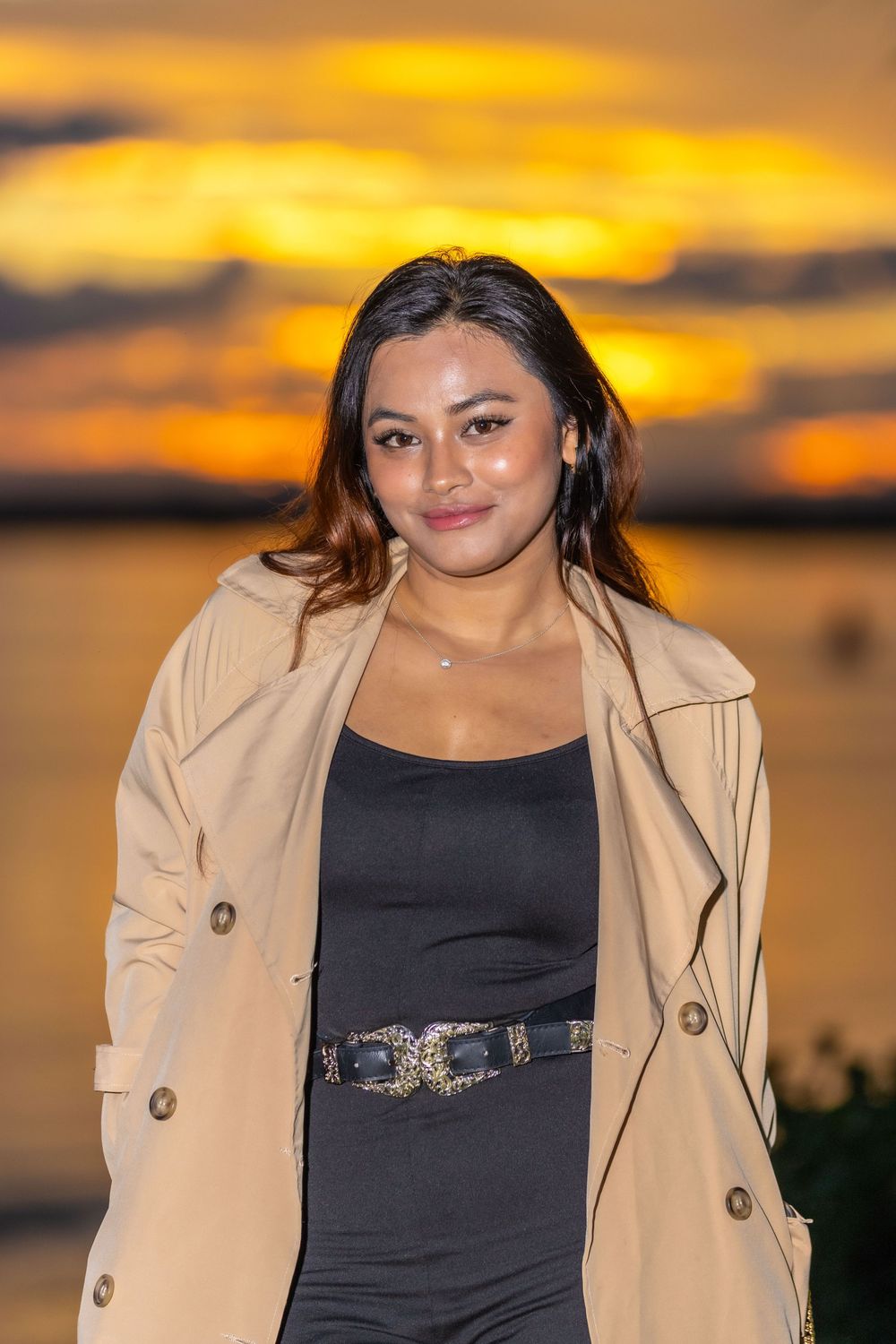 Portrait against a dramatic orange sunset sky wearing a stylish trench coat and black dress.