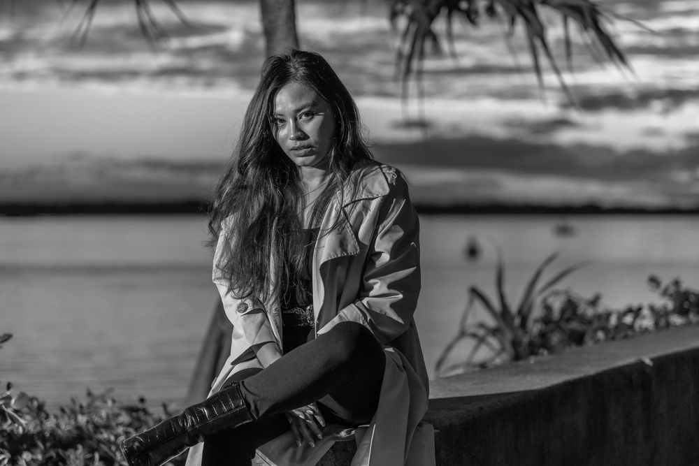Black and white portrait with scenic waterfront backdrop features stylish outerwear and casual pose.
