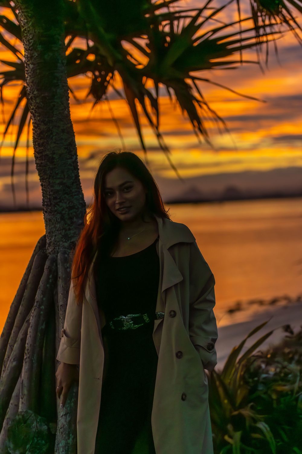 A silhouette stands by a palm tree against a vibrant orange sunset sky over a tropical beach.