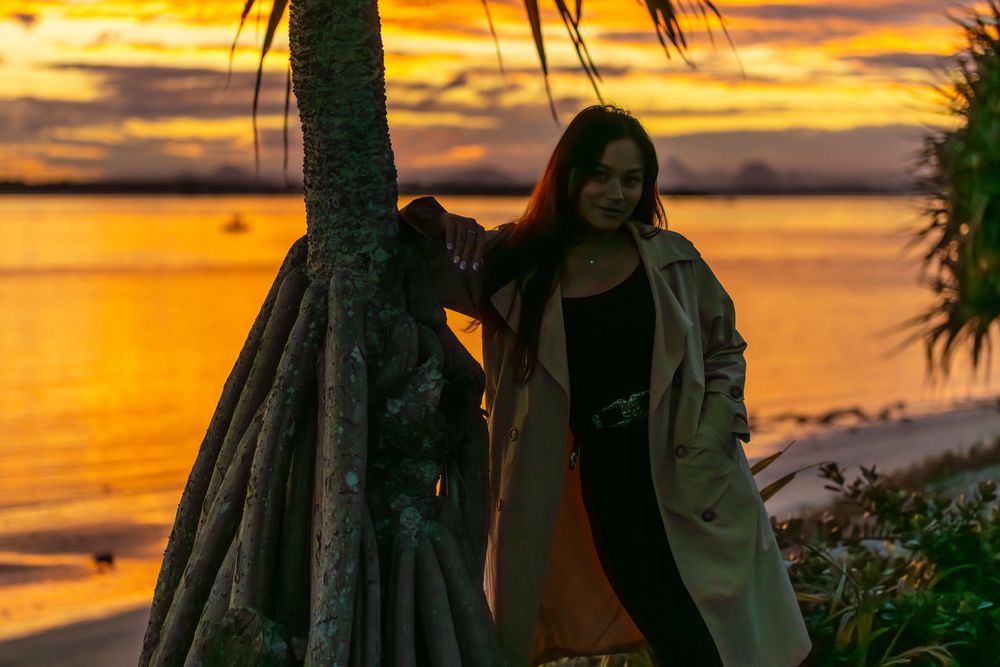 Figure stands by a palm tree silhouetted against a vibrant orange sunset over the ocean.