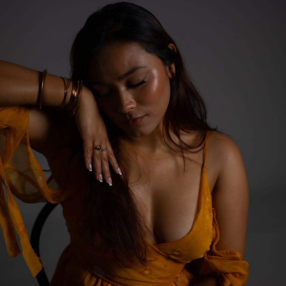 Model poses gracefully in mustard yellow outfit against dark background with dramatic lighting and shadows.