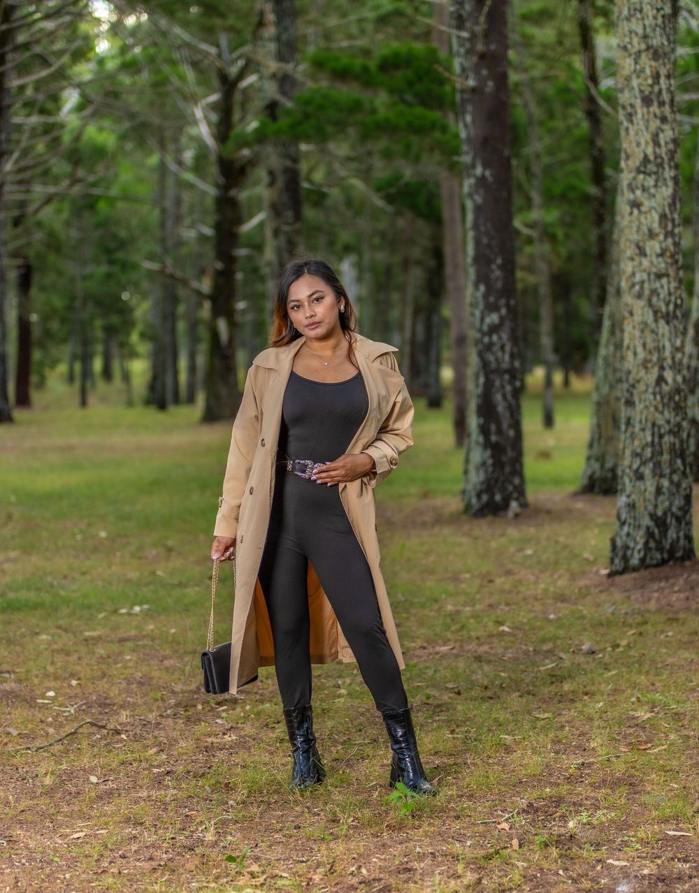 Figure in black clothing and tan jacket poses among tall pine trees in a forest setting.