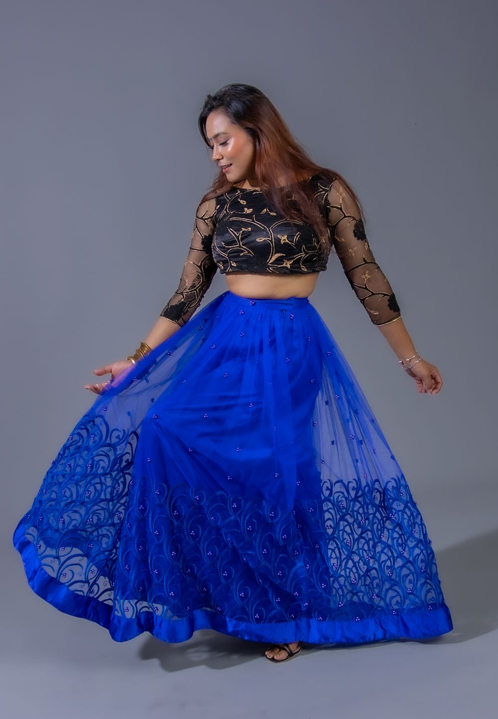 Dancing pose in a black mesh top and vibrant blue floor-length skirt with intricate pattern detailing.