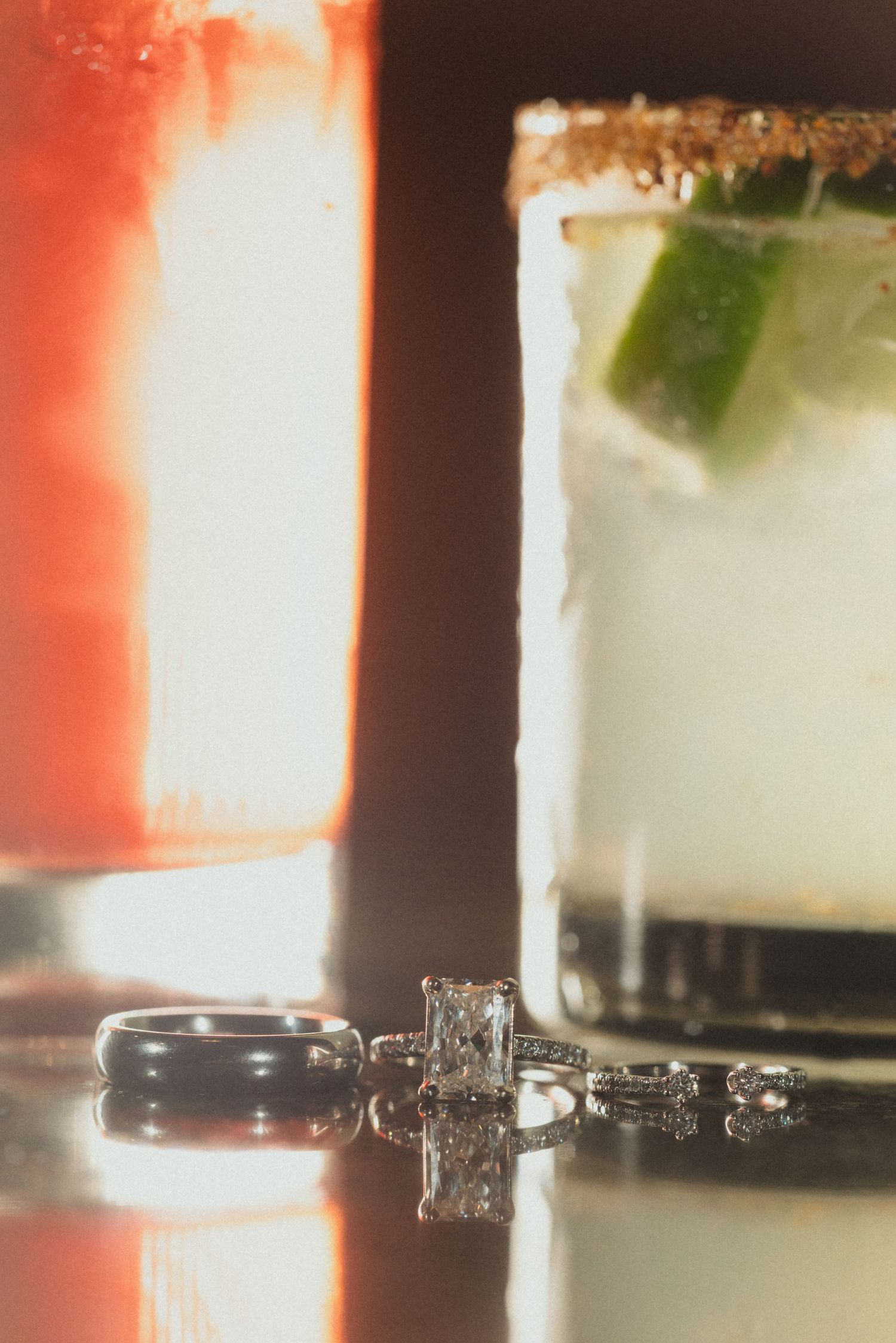 Wedding rings reflected on glossy surface beside colorful cocktails at a wedding reception.