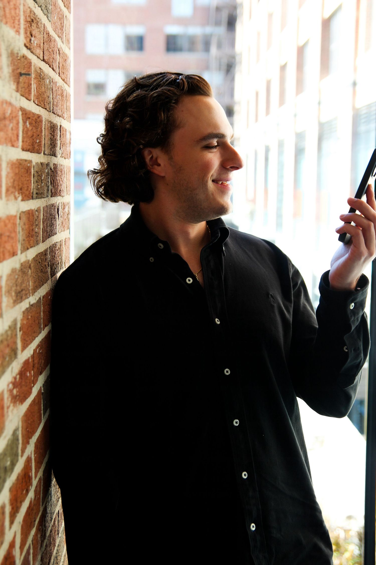A person in a black button-down shirt smiles while leaning against a brick wall near a window.