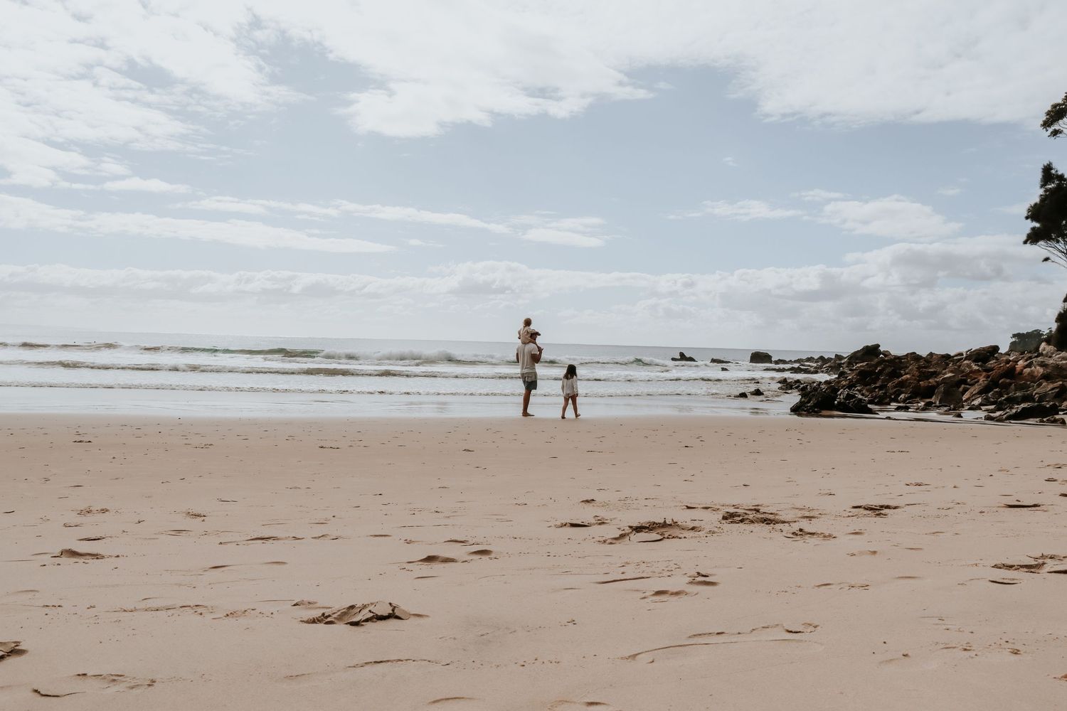 Adult and child walking on a sandy beach near rocky shores with gentle ocean waves under a cloudy sky.
