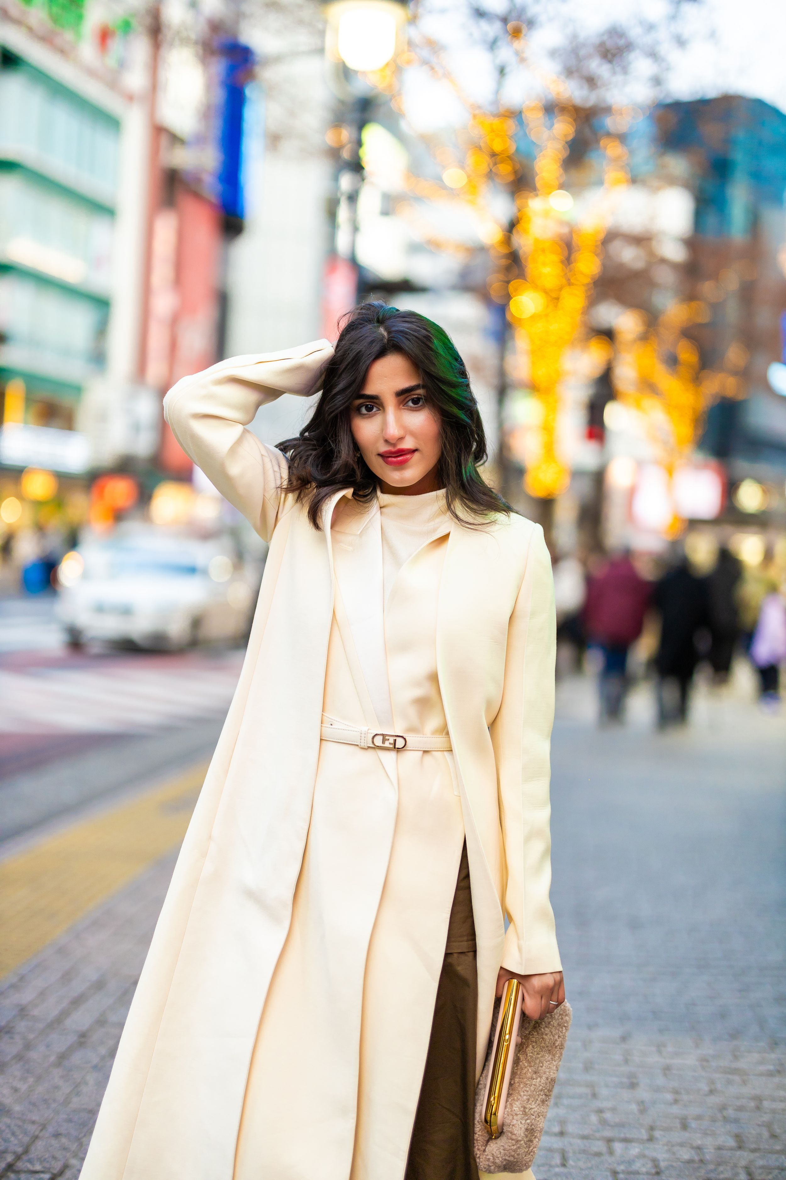 A stylish woman in a cream coat poses on a city street with colorful lights and buildings behind her.