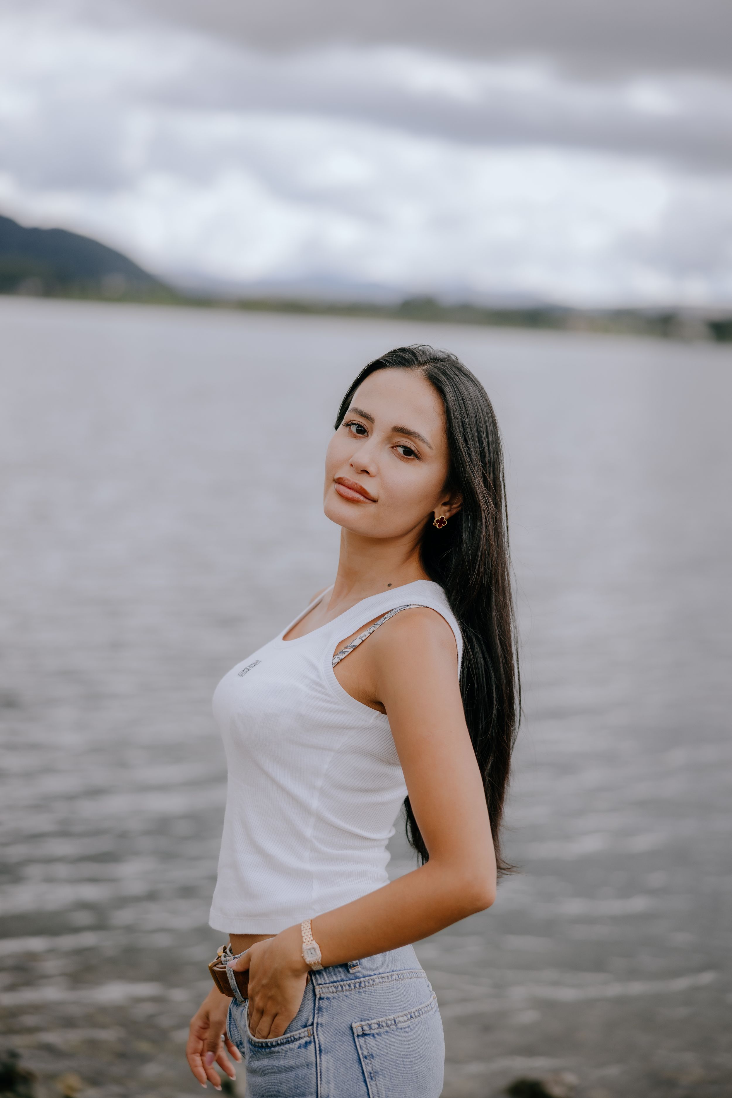 Person in white tank top and jeans poses against cloudy sky and water background.
