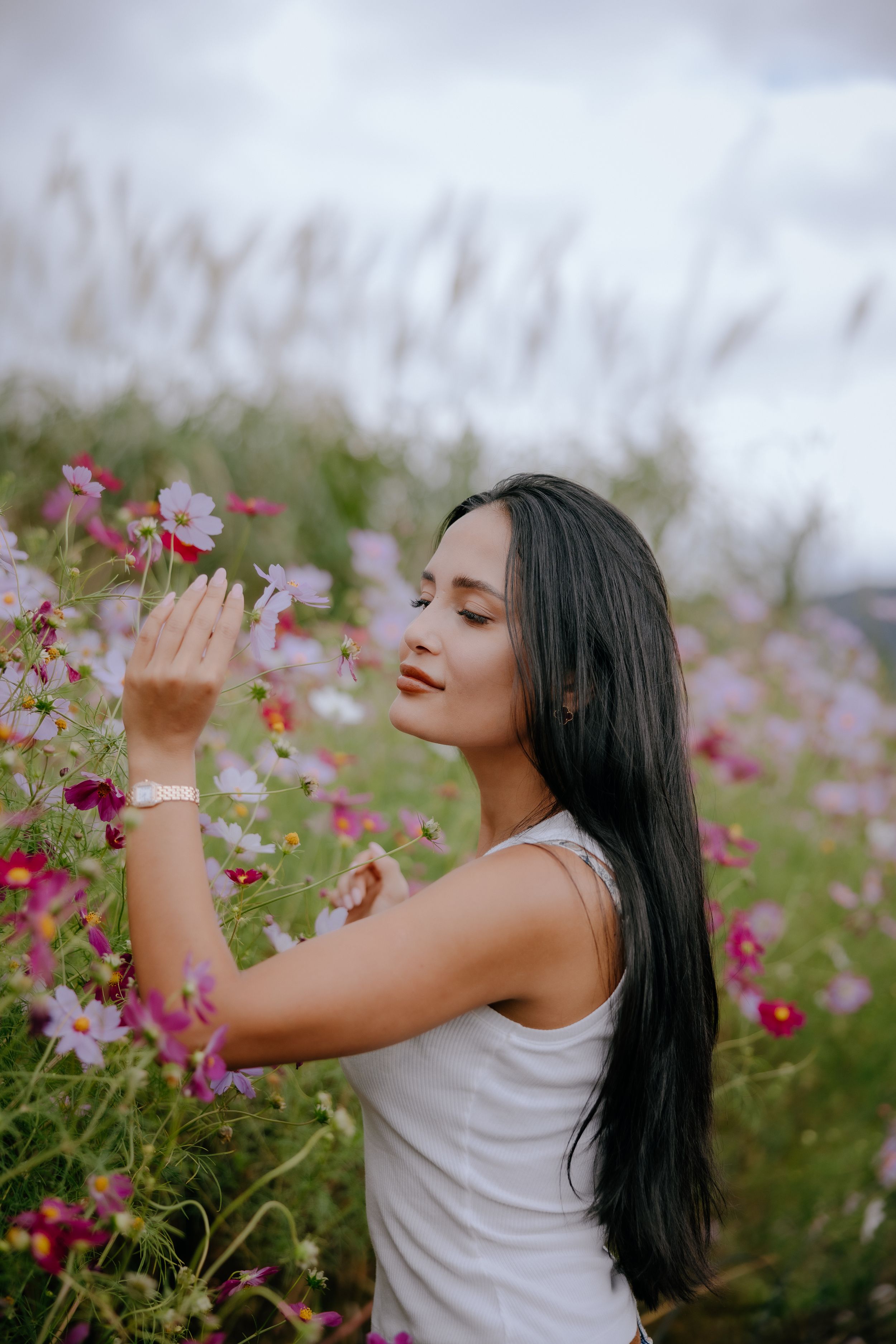 A person in a white tank top poses joyfully in a field of pink cosmos flowers on a cloudy day.