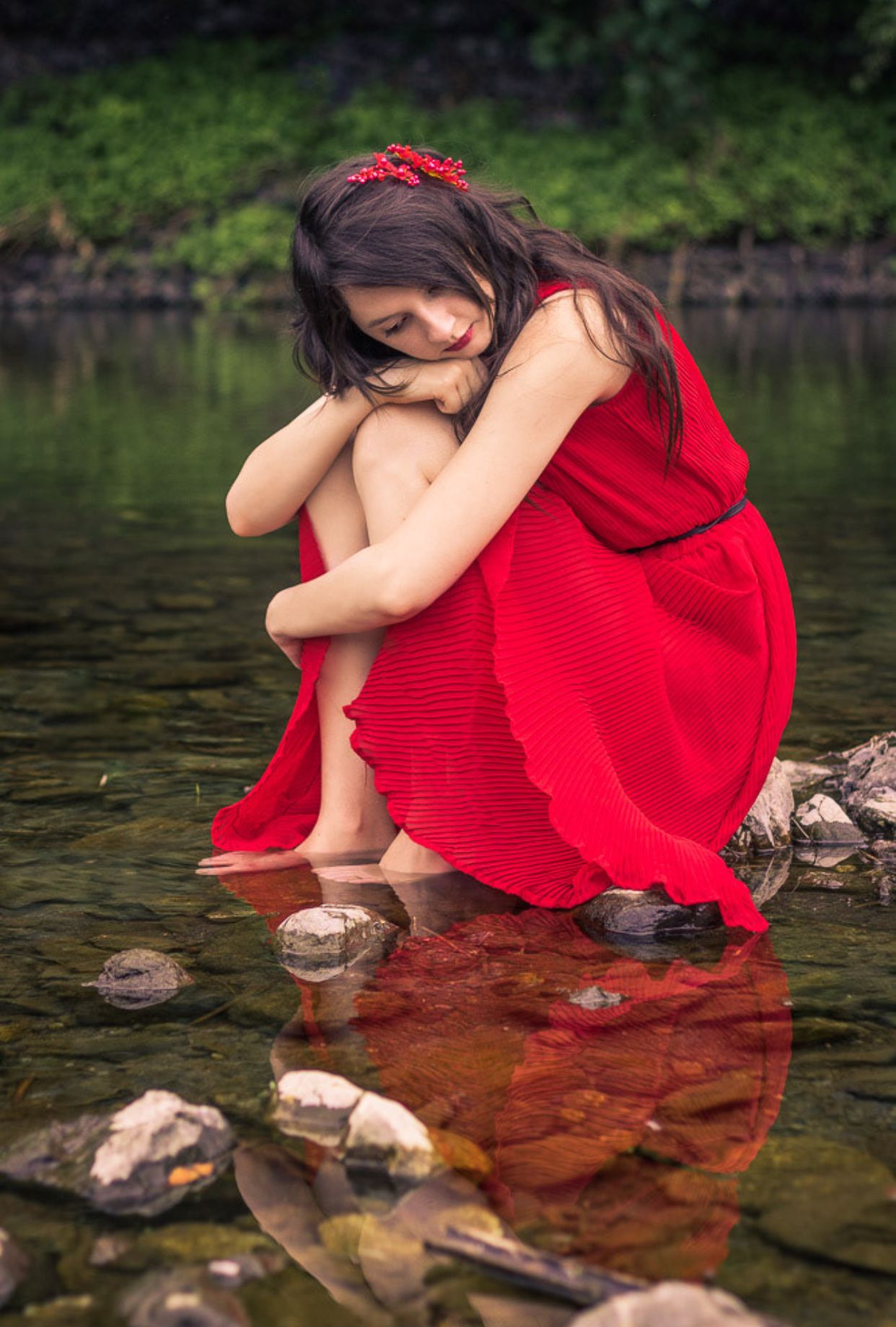 Contemplative pose in red dress sitting by tranquil stream with stones.