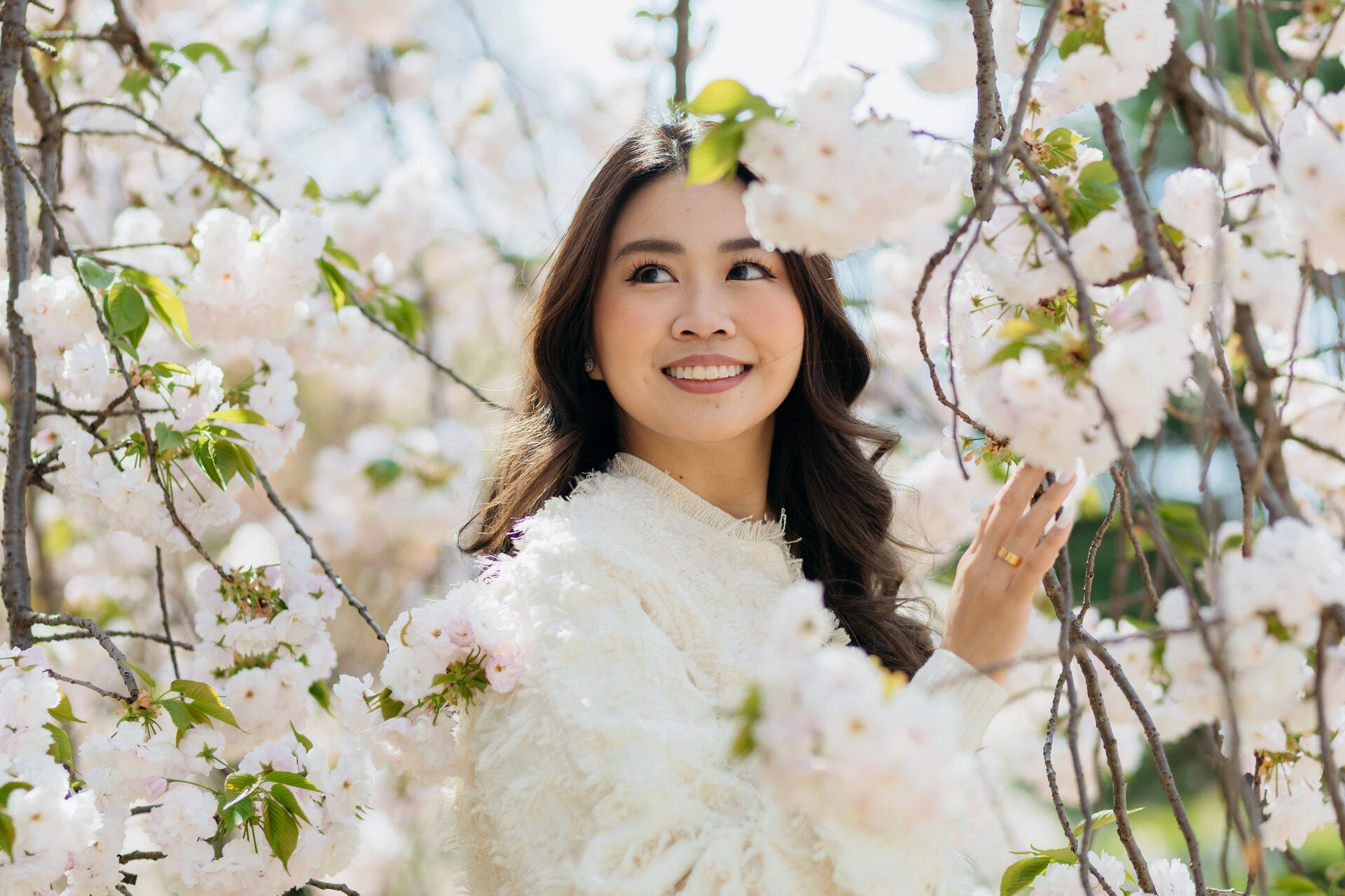 Woman in white sweater smiling among cherry blossom branches.