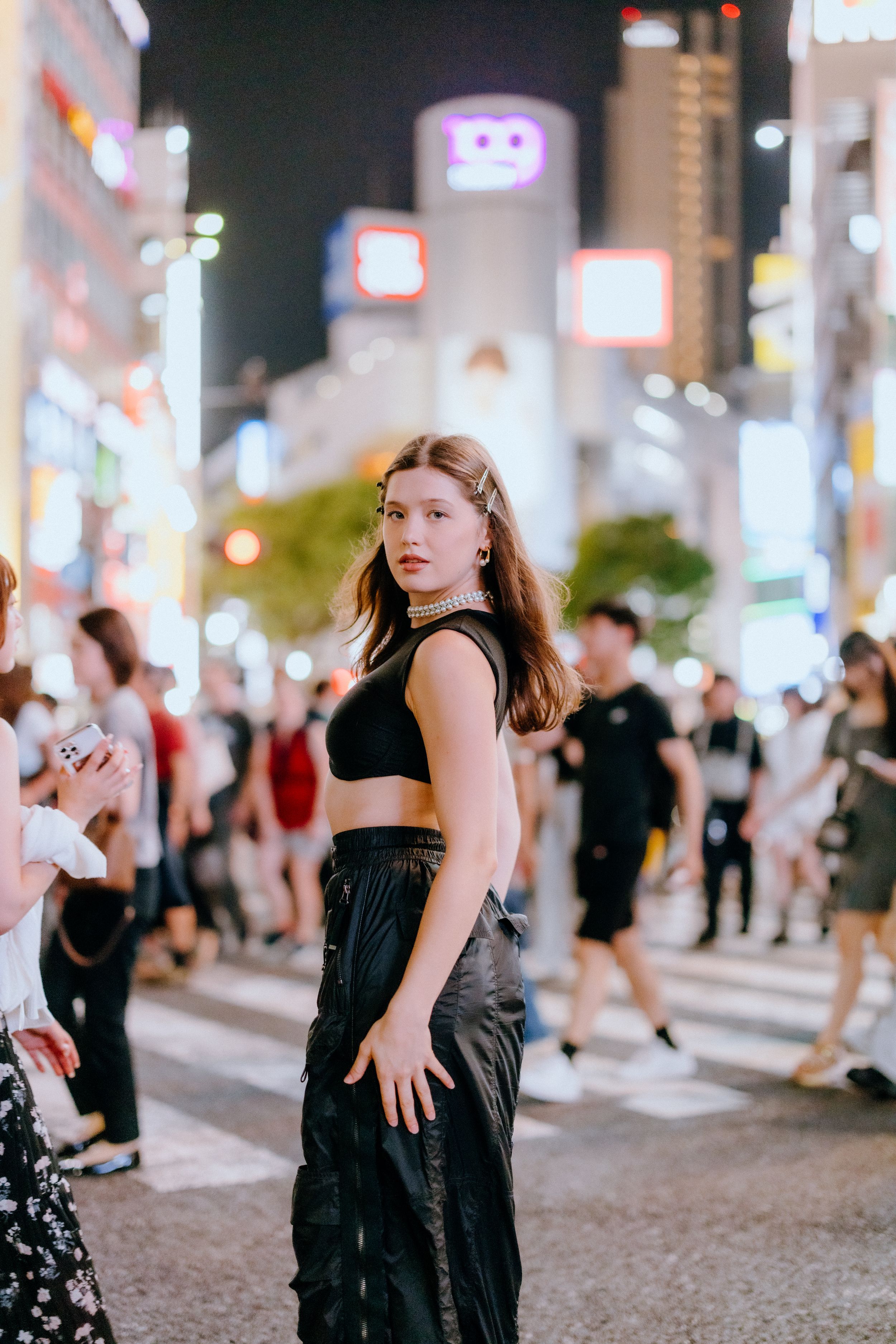 Street fashion photo at night in Tokyo's Shibuya district with neon lights and crowds in the background.