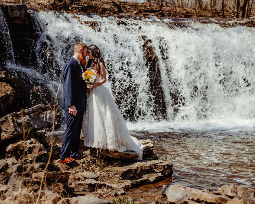 A romantic wedding photo captured in front of a cascading waterfall with rocks and natural scenery.