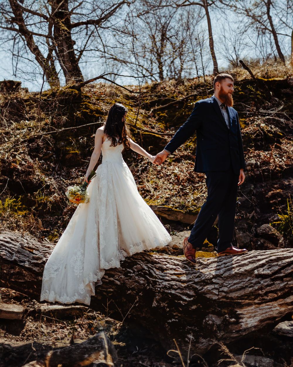 A couple in wedding attire balancing on a fallen tree trunk in a rustic forest setting.