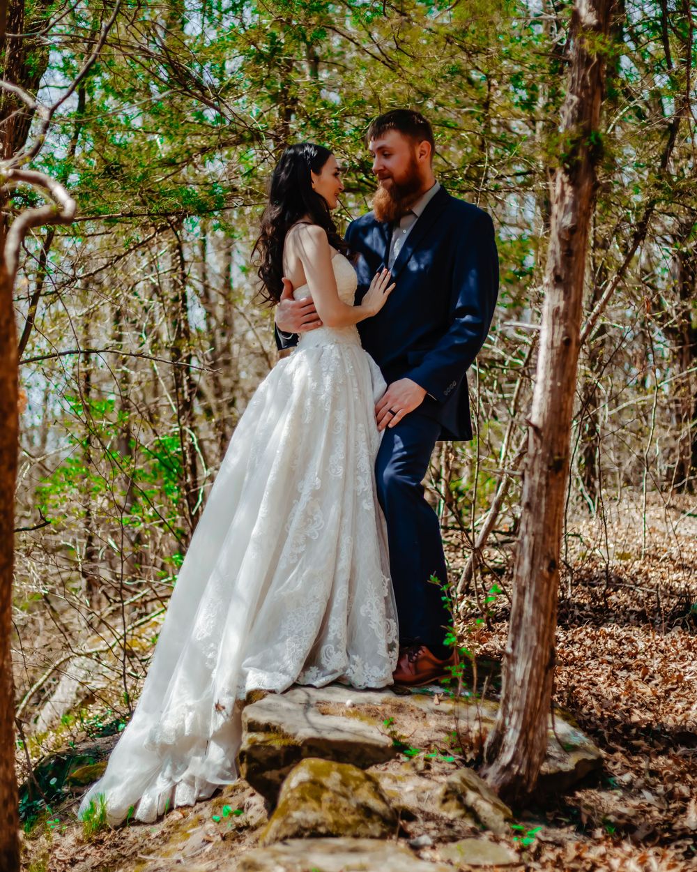 A romantic wedding moment photographed in a sunlit forest setting with natural stone elements.