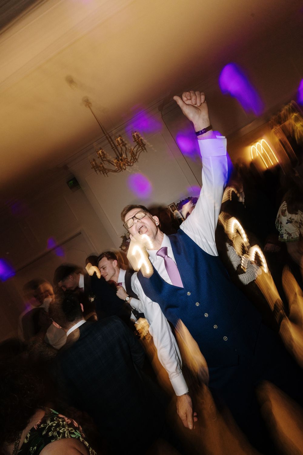 Man in navy suit celebrating on wedding dance floor with fist raised under purple lights.