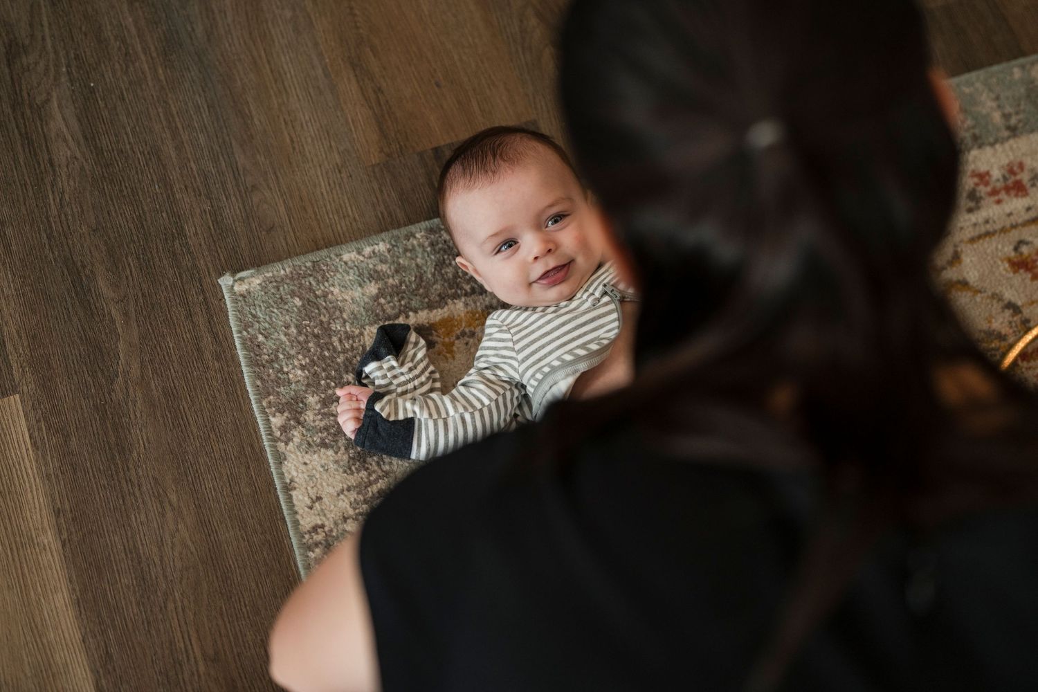 A baby in striped clothing smiles while lying on a patterned rug.